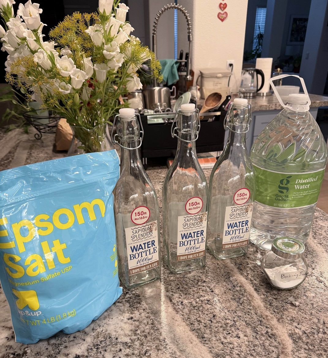 Large blue bag of epsom salt, three one-liter glass bottles with swing tops, a gallon of bottled distilled water, and a small glass container labeled 'Baking Soda', all on a kitchen countertop.